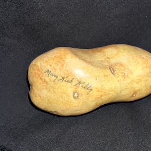 May include: A hand-painted, potato-shaped ceramic sculpture with a light brown and tan color scheme. The sculpture has a smooth texture and features the handwritten inscription "Mary Kack Kelly".