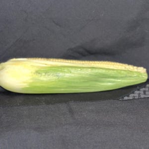 May include: An ear of corn, with a pale yellow husk and green leaves, rests on a dark surface. The corn is partially husked, revealing the kernels. The image is well-lit, highlighting the natural colors and textures of the corn.