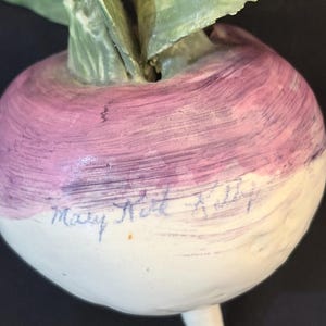 May include: A ceramic turnip sculpture with a white base and a purple top. The sculpture has green leaves and a small stem. The artist's signature, "Mary Ruth Kelly", is written in blue ink on the white base.