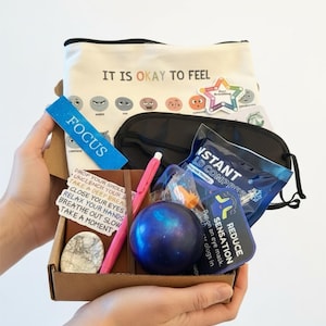 May include: A self-care gift box held in hands, containing a canvas pouch with "IT IS OKAY TO FEEL" text, a sleep mask, a blue stress ball, a pen, and other items. Includes a "FOCUS" sticker and a card with instructions.