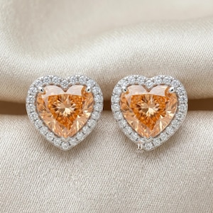 May include: A pair of heart-shaped stud earrings featuring a central orange gemstone surrounded by a halo of small white stones set in a silver-toned metal frame. The earrings have a faceted surface that reflects light, creating a bright appearance.