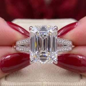 May include: A silver-toned ring featuring a large emerald-cut center stone held by four prongs. The split shank band is adorned with small pavé-set stones and features milgrain detailing along the edges. The text JJ JEWELRY BY JITA is visible.