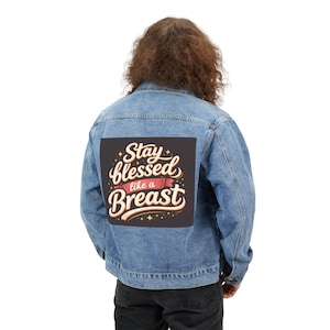 May include: A light blue denim jacket featuring a large square graphic on the back. The graphic has a dark background with the text Stay blessed like a Breast in cream-coloured cursive and block lettering, accented by a red ribbon and small stars.