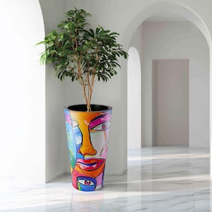 Può includere: Un'alta fioriera colorata con un design di volto astratto dipinto. La fioriera contiene un piccolo albero con foglie verdi e un tronco marrone. Il design della fioriera presenta tonalità arancioni, blu, rosa e rosse. La fioriera si trova in un corridoio bianco con porte ad arco.