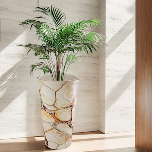 Può includere: Un vaso da fiori alto e decorativo con un design unico in pietra screpolata nei toni del bianco, beige e marrone. Il vaso contiene una lussureggiante pianta di palma verde. Il vaso è all'interno, vicino a una porta di legno e a un muro di piastrelle bianche.