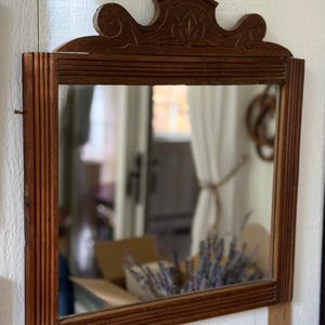 May include: A rectangular wall mirror with a medium-brown wooden frame featuring vertical fluted side panels and a decorative carved crest at the top. The mirror glass is clear and reflects the surrounding indoor environment.
