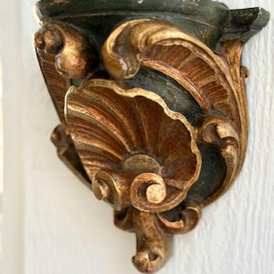May include: A wall-mounted shelf bracket featuring a dark green finish with gold-toned highlights. The piece displays an intricate carved design with a central shell motif, scrolling foliage, and a flat top surface for display purposes.