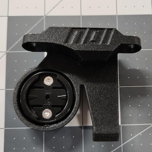 Pode incluir: Suporte preto para computador de bicicleta com base circular e dois parafusos prateados. O suporte tem uma superf&iacute;cie texturizada e uma extens&atilde;o retangular. Projetado para segurar um computador de ciclismo.