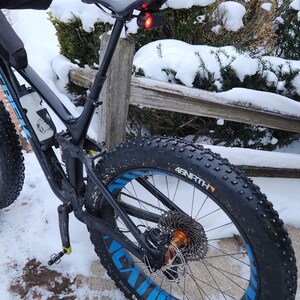 Pode incluir: Uma bicicleta preta de pneus largos com detalhes azuis nas rodas est&aacute; estacionada na neve. Os pneus exibem as palavras "Dillinger" e "45NRTH". Uma luz traseira vermelha est&aacute; montada no espig&atilde;o do selim.