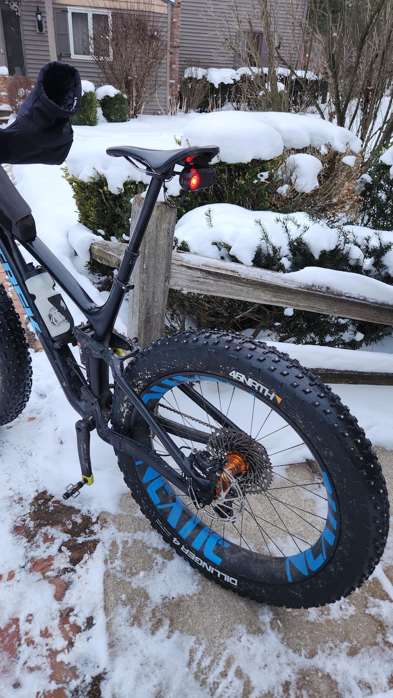 Pode incluir: Uma bicicleta preta de pneus largos com detalhes azuis nas rodas est&aacute; estacionada na neve. Os pneus exibem as palavras "Dillinger" e "45NRTH". Uma luz traseira vermelha est&aacute; montada no espig&atilde;o do selim.