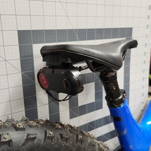 Pode incluir: Assento de bicicleta preto com uma luz traseira vermelha e uma c&acirc;mera preta acoplada. A bicicleta tem um quadro azul e um pneu preto. O assento &eacute; rotulado como "BODY GEOMETRY". O pneu est&aacute; coberto de sujeira.