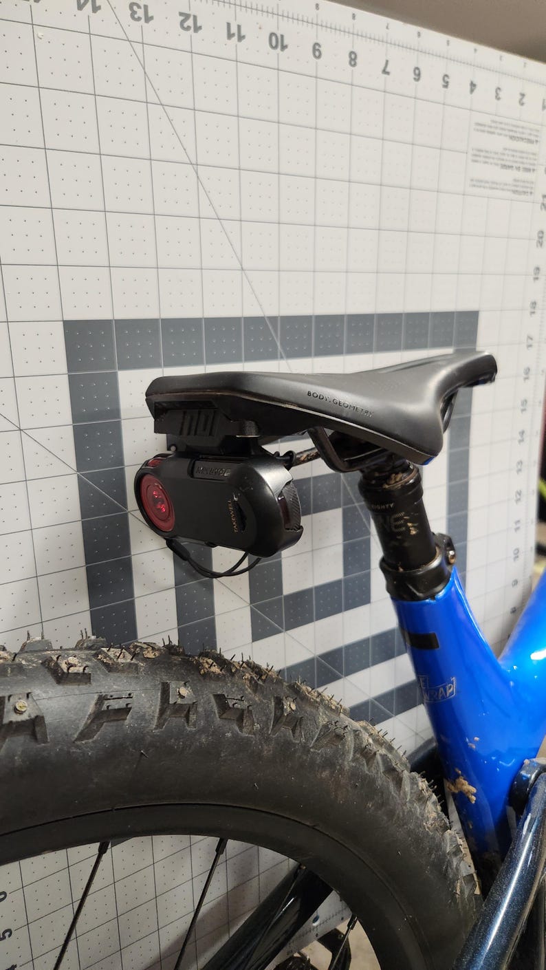 Pode incluir: Assento de bicicleta preto com uma luz traseira vermelha e uma c&acirc;mera preta acoplada. A bicicleta tem um quadro azul e um pneu preto. O assento &eacute; rotulado como "BODY GEOMETRY". O pneu est&aacute; coberto de sujeira.