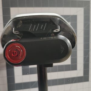 Pode incluir: Um selim de bicicleta preto com um dispositivo Garmin preto ligado. O dispositivo tem uma luz vermelha e um corpo preto. O selim &eacute; montado num poste preto. O fundo &eacute; uma grelha cinzenta e branca.