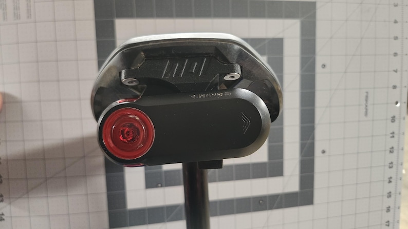 Pode incluir: Um selim de bicicleta preto com um dispositivo Garmin preto ligado. O dispositivo tem uma luz vermelha e um corpo preto. O selim &eacute; montado num poste preto. O fundo &eacute; uma grelha cinzenta e branca.