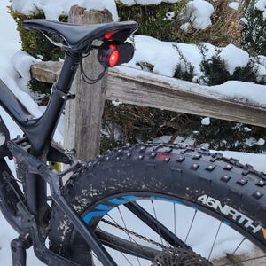 Pode incluir: Uma bicicleta de montanha preta com um assento preto e uma luz traseira vermelha. A bicicleta tem um pneu grosso com detalhes em azul e o nome da marca "45NRTH" no pneu. O fundo mostra arbustos cobertos de neve e uma cerca de madeira.