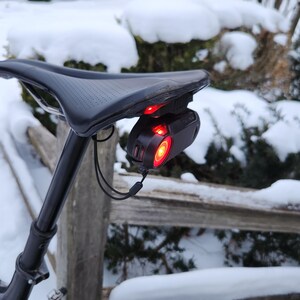 Pode incluir: Selim de bicicleta preto com uma luz traseira vermelha presa. A luz traseira &eacute; preta e tem uma luz vermelha brilhante. A bicicleta est&aacute; ao ar livre em um ambiente com neve.