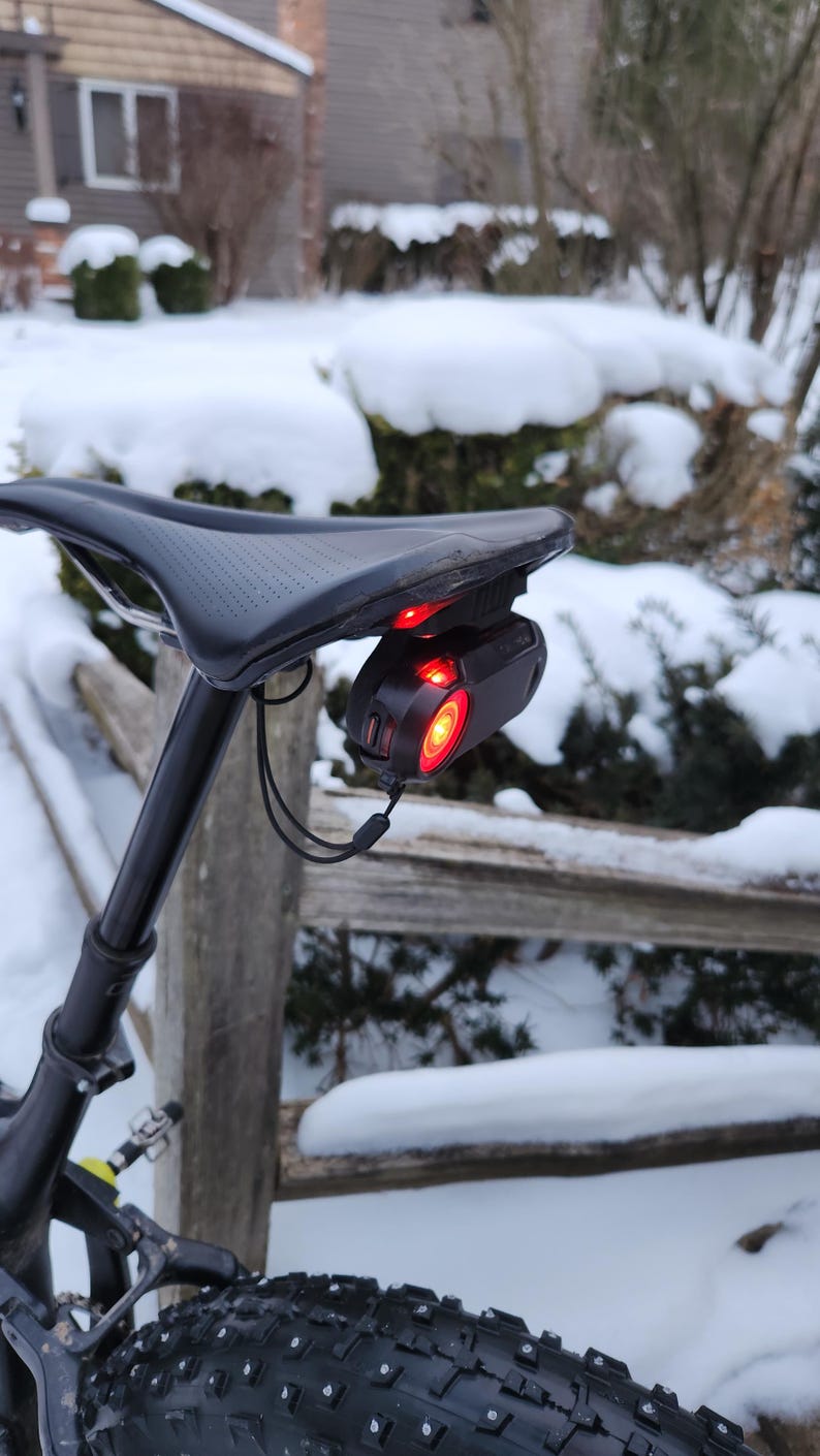 Pode incluir: Selim de bicicleta preto com uma luz traseira vermelha presa. A luz traseira &eacute; preta e tem uma luz vermelha brilhante. A bicicleta est&aacute; ao ar livre em um ambiente com neve.