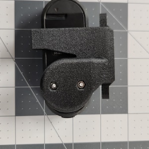 Pode incluir: Um dispositivo de pl&aacute;stico preto com uma superf&iacute;cie texturizada, apresentando uma base arredondada e uma extens&atilde;o retangular. Dois rebites prateados s&atilde;o vis&iacute;veis na se&ccedil;&atilde;o arredondada. O dispositivo est&aacute; sobre um fundo quadriculado cinza e branco.