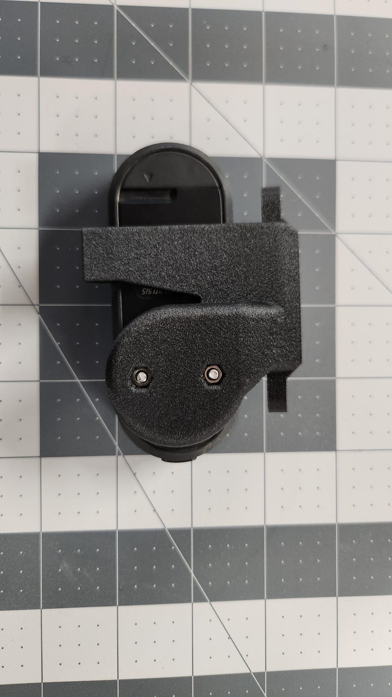 Pode incluir: Um dispositivo de pl&aacute;stico preto com uma superf&iacute;cie texturizada, apresentando uma base arredondada e uma extens&atilde;o retangular. Dois rebites prateados s&atilde;o vis&iacute;veis na se&ccedil;&atilde;o arredondada. O dispositivo est&aacute; sobre um fundo quadriculado cinza e branco.