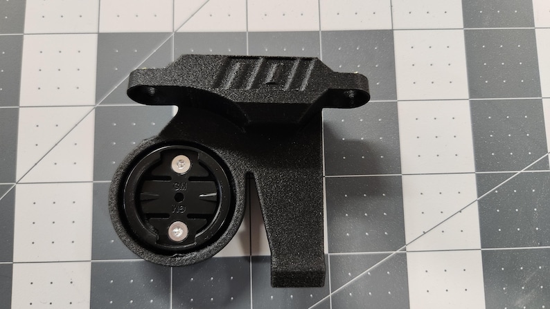 Pode incluir: Suporte preto para computador de bicicleta com base circular e dois parafusos prateados. O suporte tem uma superf&iacute;cie texturizada e uma extens&atilde;o retangular. Projetado para segurar um computador de ciclismo.