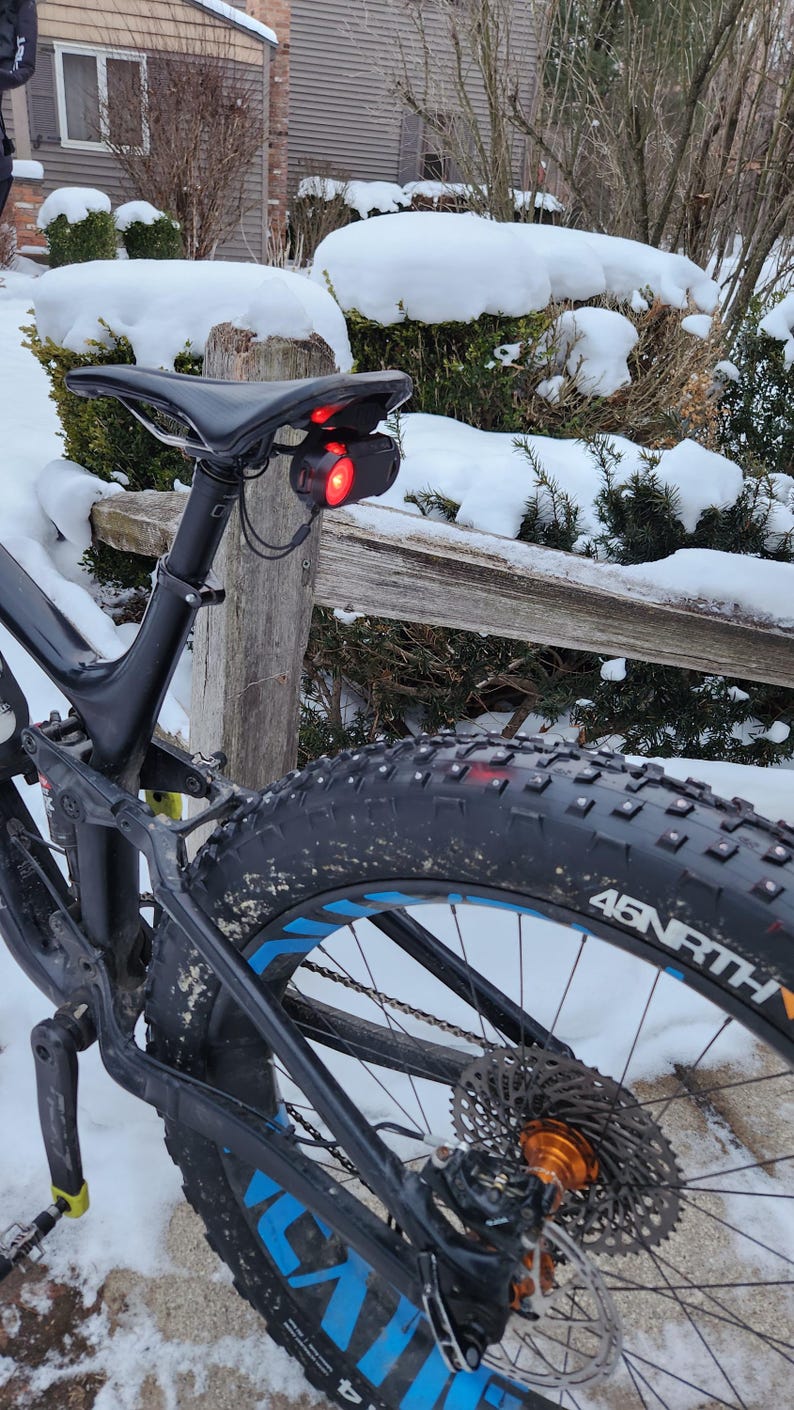 Pode incluir: Uma bicicleta de montanha preta com um assento preto e uma luz traseira vermelha. A bicicleta tem um pneu grosso com detalhes em azul e o nome da marca "45NRTH" no pneu. O fundo mostra arbustos cobertos de neve e uma cerca de madeira.