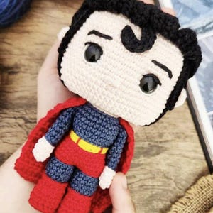 Puede incluir: Muñeco de Superman hecho a mano con ganchillo. El muñeco tiene una cara color crema con pelo negro, un traje azul y rojo y un cinturón amarillo. El muñeco tiene una capa roja y está en manos de alguien.