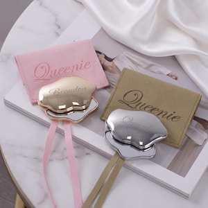 Peut inclure: Deux miroirs de poche aux formes irrégulières avec des rubans, l'un doré avec le nom Brandon et l'autre argenté avec le nom Queenie, chacun accompagné d'une pochette en tissu doux portant le nom brodé Queenie.