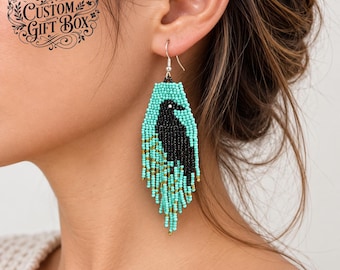 Pode incluir: Brincos pendentes com uma silhueta de pássaro preto feita de pequenas contas sobre um fundo de contas turquesa. O design inclui uma franja de contas turquesa e douradas na parte inferior. O texto The Quincy Archive Custom Gift Box é visível.