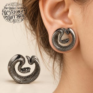 Puede incluir: Una dilatación de oreja plateada con un diseño de serpiente enroscada con escamas detalladas. La joya circular tiene un centro abierto donde descansa la cabeza de la serpiente. El texto "The Quincy Archive Custom Gift Box" aparece arriba a la izquierda.