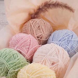 May include: A collection of six round balls of soft, velvet-textured yarn in pastel shades of light green, pale yellow, soft pink, dusty rose, beige, and light blue, arranged together inside a light peach-colored tissue paper wrapping.