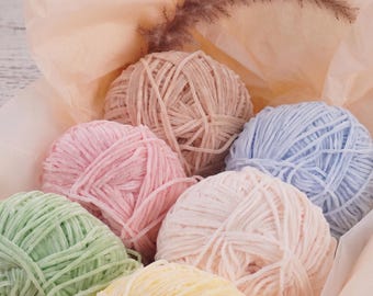 May include: A collection of six round balls of soft, velvet-textured yarn in pastel shades of light green, pale yellow, soft pink, dusty rose, beige, and light blue, arranged together inside a light peach-coloured tissue paper wrapping.