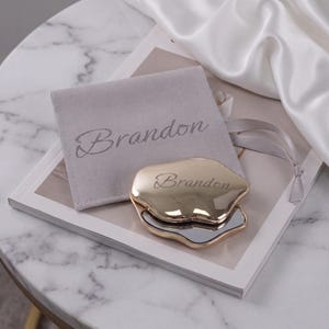 May include: A gold-toned, irregular-shaped compact mirror featuring the name Brandon engraved on the surface. It is accompanied by a light gray velvet pouch with the name Brandon printed in a script font on the front.