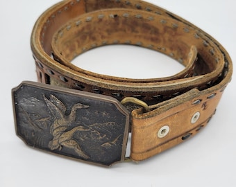 May include: A tan leather belt featuring a rectangular metal buckle with a dark, embossed relief of two flying ducks over a landscape. The belt strap has decorative stitching along the edges and two silver-toned metal grommets near the buckle.