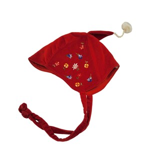 May include: A red velvet bonnet featuring embroidered floral patterns in yellow, blue, and white. The headwear includes a pointed top with a small white pom-pom attached by a cord, and two long red velvet ties extending from the bottom edges.