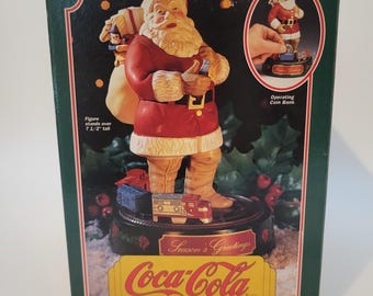 May include: A Coca-Cola Santa Claus mechanical coin bank in a green box. The box features a graphic of Santa holding a bottle, a toy sack, and a toy truck on a base. Text includes "Season's Greetings", "Coca-Cola", and "Figure stands over 7 1/2" tall".