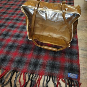 May include: A brown vinyl tote bag with a clear front panel, white piping, and two shoulder straps. The bag is placed on a red, black, and gray plaid wool blanket with fringe edges and a blue Pendleton brand label in the bottom corner.