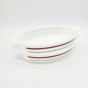 May include: Two stacked oval-shaped white ceramic baking dishes featuring two thin, dark red horizontal stripes running along the exterior sides. Each dish includes small integrated handles on both ends for easy gripping and handling.
