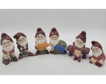 May include: A set of six ceramic gnome figurines, each wearing a maroon pointed hat. The gnomes are depicted in various poses, including holding a yellow watering can, sitting on a brown bench, and resting, all featuring white beards and colorful clothing.