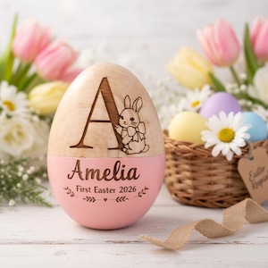 May include: A wooden Easter egg with a pink base, engraved with the name "Amelia" and "First Easter 2026." The top half features a large letter "A" and a bunny illustration. A basket of colorful eggs and flowers are in the background.