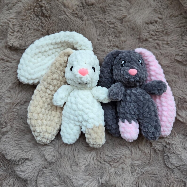 Pu&ograve; includere: Due peluche a forma di coniglietto. Uno &egrave; bianco con dettagli beige e rosa, l'altro &egrave; grigio con dettagli rosa. Entrambi hanno il naso rosa e sono realizzati in un materiale morbido e strutturato. I coniglietti sono su una superficie marrone strutturata.