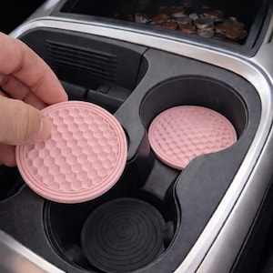 Puede incluir: Posavasos rosas para portavasos de coche con un patrón de panal. Uno está siendo sostenido por una mano, otro está en el portavasos. Un posavasos negro está en el otro portavasos. El interior del coche es visible.