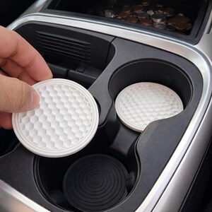 May include: White, round car coasters with a golf ball pattern. One coaster is held by a hand, while another sits in a car cup holder. A black coaster with concentric circles is also visible in the cup holder.