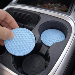 May include: Light blue car cup holder coasters with a golf ball pattern. One coaster is being held by a hand, while another sits in a cup holder. The car's interior is visible in the background.