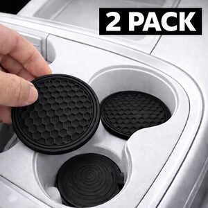 May include: Black car coasters with a honeycomb pattern are shown, with one being held by a hand. The coasters are round and fit into a car's cup holder. The text "2 PACK" is in a black rectangle in the top right corner.