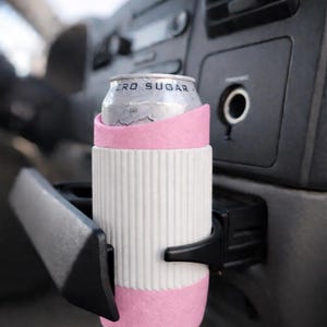 May include: A white and pink drink insulator with a can of soda inside, held in a car's cup holder. The can is silver with the words "ZERO SUGAR" printed on it. The insulator has a ribbed design.