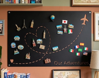 Puede incluir: Un tablero magnético negro rectangular con una forma de corazón cortada con láser y una trayectoria de vuelo discontinua que termina en una silueta de avión. El tablero muestra el texto Our Adventures y varios imanes de viaje.