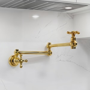 May include: A gold-colored, wall-mounted pot filler faucet with a cross-handle design. The faucet has a double-jointed arm for extending over a cooktop. The fixture is mounted on a white marble-patterned wall.
