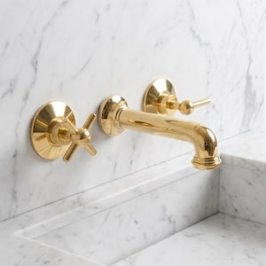 May include: A gold-colored bathroom faucet with a cross-shaped handle and a spout. The faucet is mounted on a white marble wall above a matching marble sink. The design is modern and luxurious.