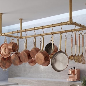 May include: A ceiling-mounted brass pot rack featuring a double-bar design with multiple S-hooks. Hanging from the hooks are various copper-toned cookware pieces, including saucepans, lids, a skillet, a ladle, a slotted spoon, and a wire whisk.