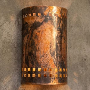 May include: Cylindrical copper wall sconce with a mottled, dark finish. The sconce has square cutouts at the top and bottom, allowing light to filter through. The fixture is mounted on a textured, gray wall.